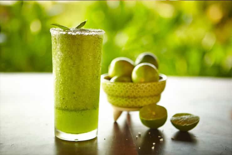 A glass of green juice next to a bowl of limes on a table.