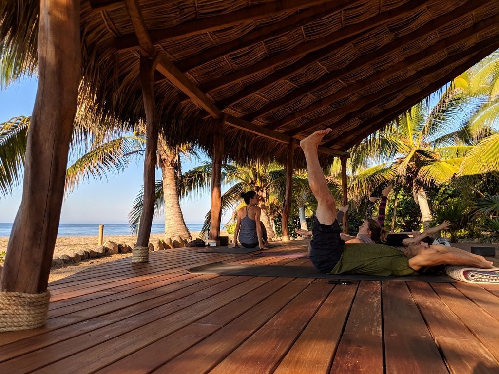A group of people are doing yoga on a wooden deck under a thatched roof.