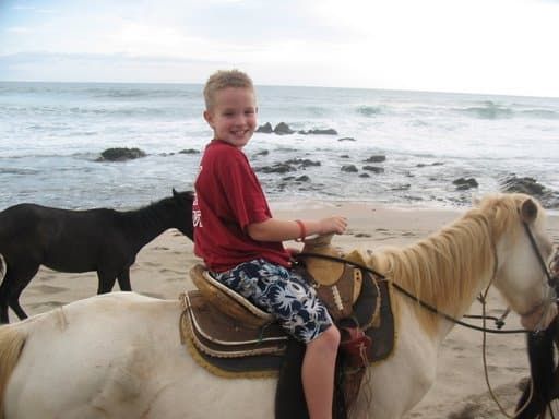 A boy in a red shirt is riding a horse on the beach