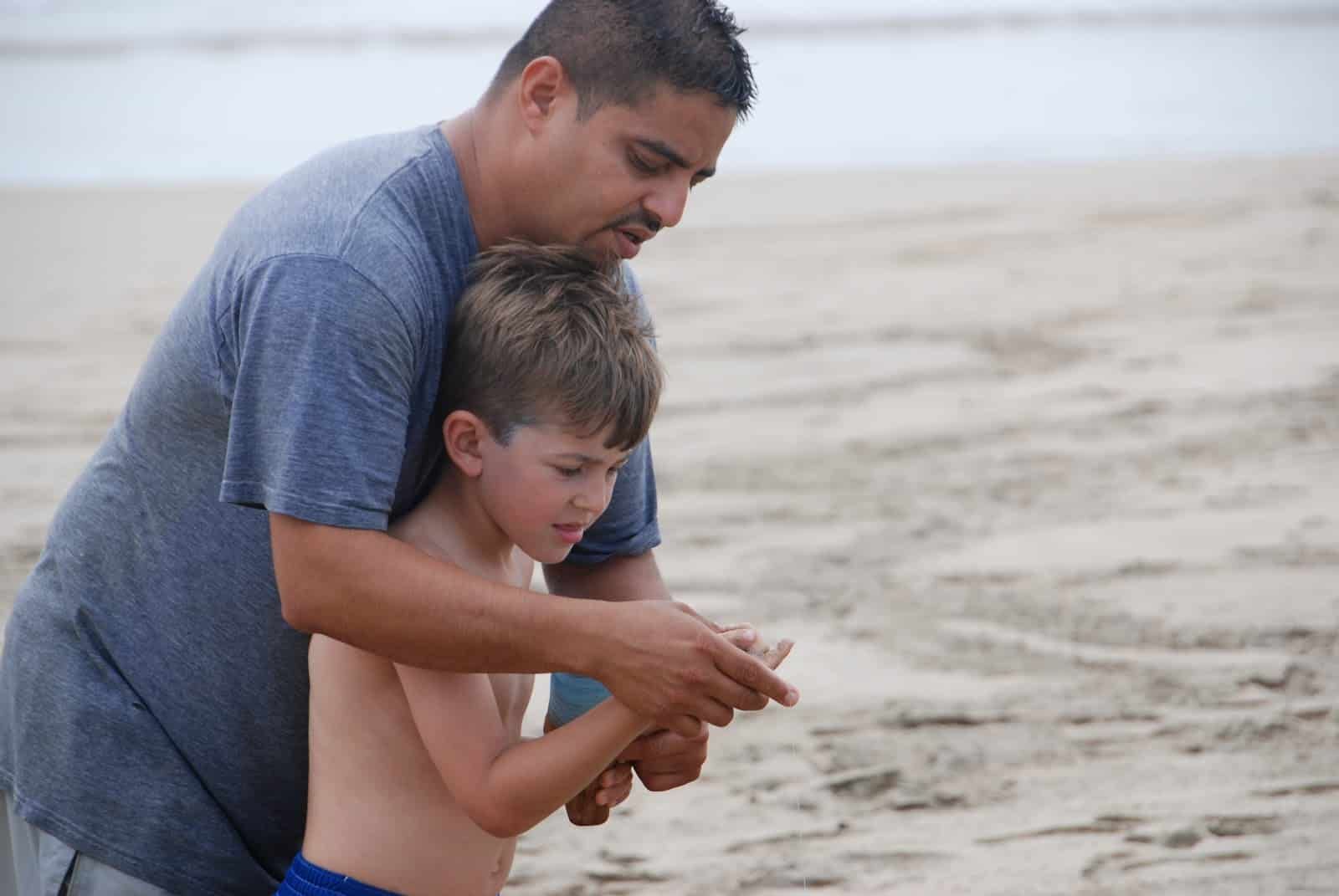 A man and a boy are standing on a beach looking at something.