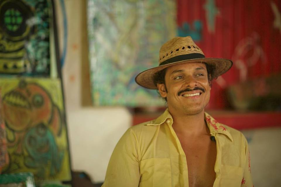 A man wearing a hat and a yellow shirt is smiling.