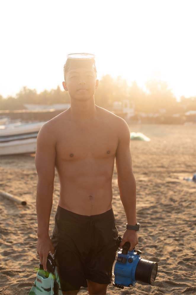 A shirtless man is standing on a beach holding a camera.