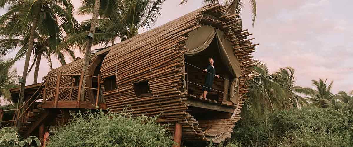 A man is standing on a balcony of a tree house surrounded by palm trees.