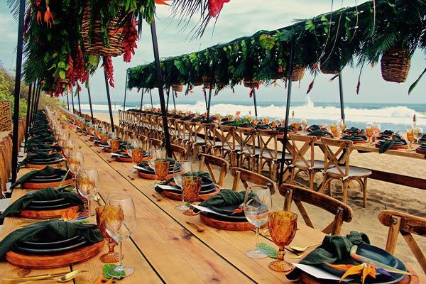 A long wooden table with plates , glasses , and napkins on it on the beach.