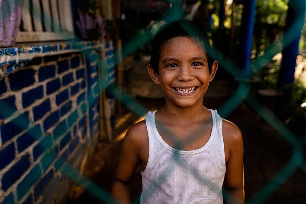 A young boy in a white tank top is smiling behind a fence.