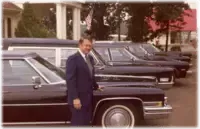 Man in a suit standing next to three black hearses outside a building.