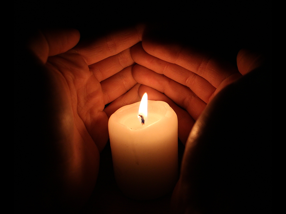 Hands cupping a lit white candle in a dark setting, providing protection and warmth.
