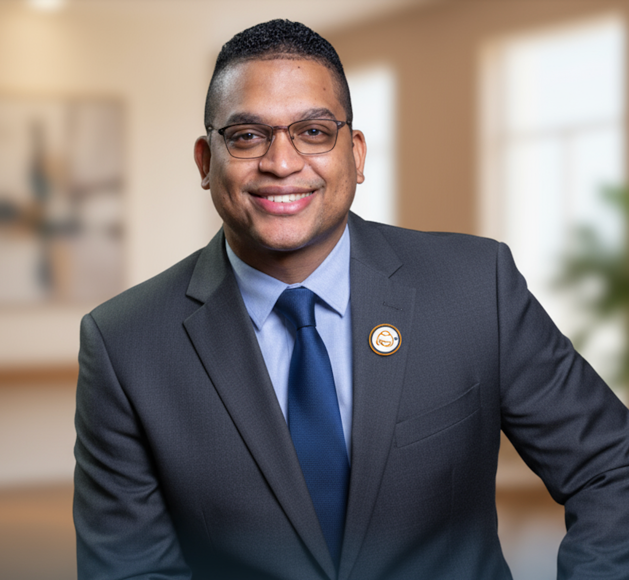 Un hombre de traje sonríe y lleva gafas. Interior con fondo neutro.