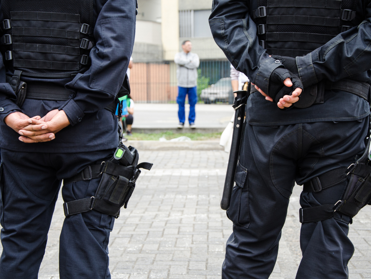 Two armed police officers in uniform stand guard. A man in casual clothes stands in the distance.
