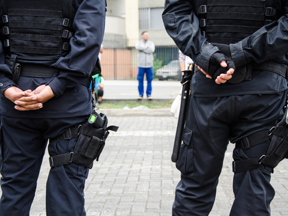 Two police officers in tactical gear stand guard; a man stands in the distance with his arms crossed.