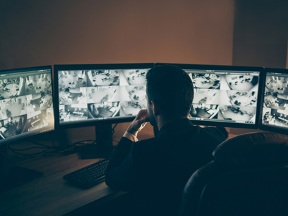 Man monitors multiple screens showing surveillance footage in a dimly lit room.