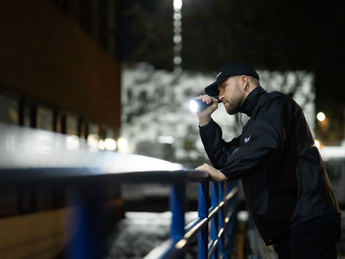 Security guard in black jacket, holding a flashlight, looks over a blue railing at night.