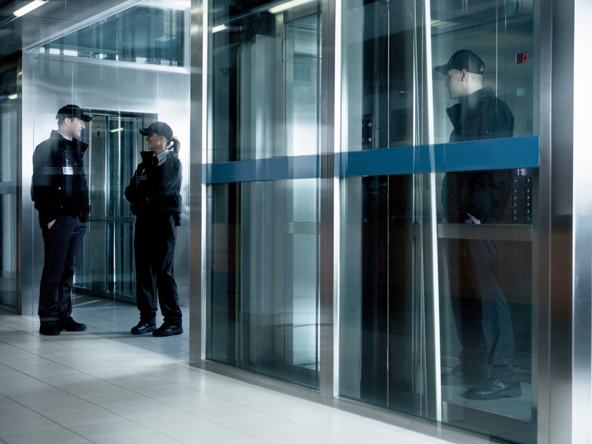 Security guards in a building hallway, near elevators. One guard inside, two outside, conversing.