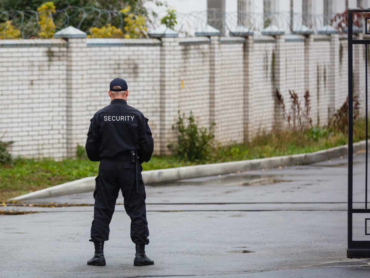 Security guard in black uniform stands watch by gate, brick wall with barbed wire in background.