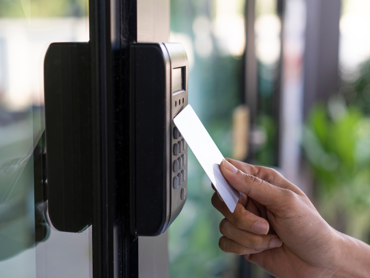 Hand swiping a white access card on a black door reader to open a glass door.