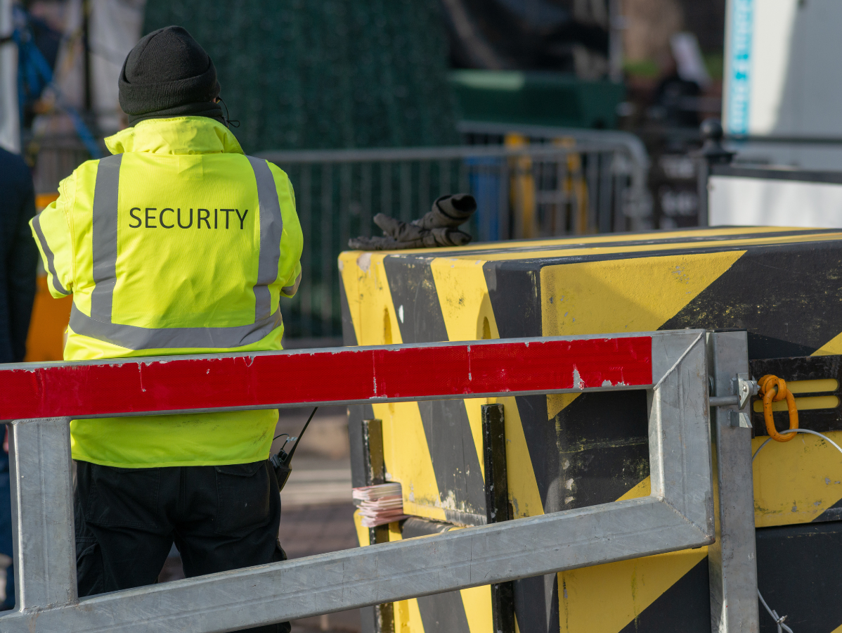 Security guard in yellow vest stands near barrier, with black and yellow blocks.