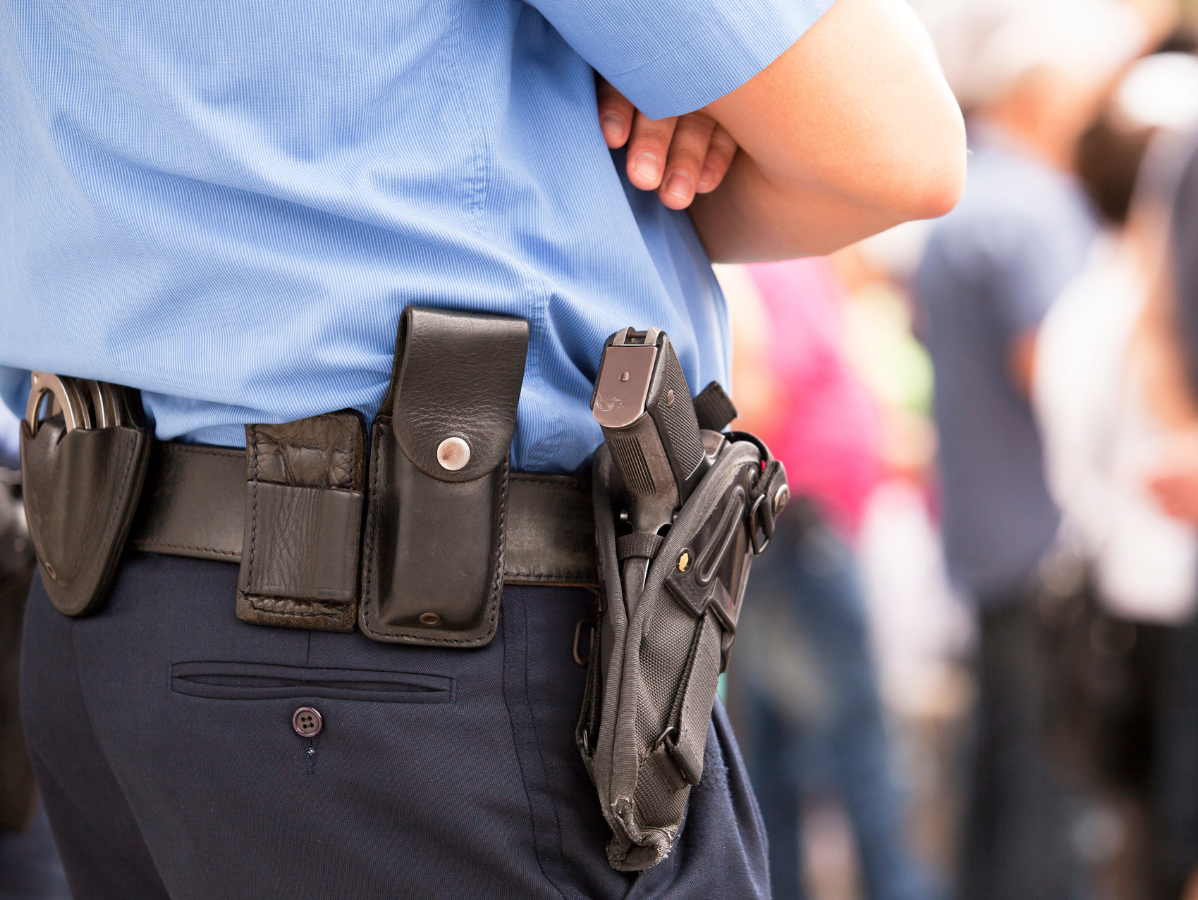 Police officer in uniform, side view, with gun and equipment on belt, watching a crowd.