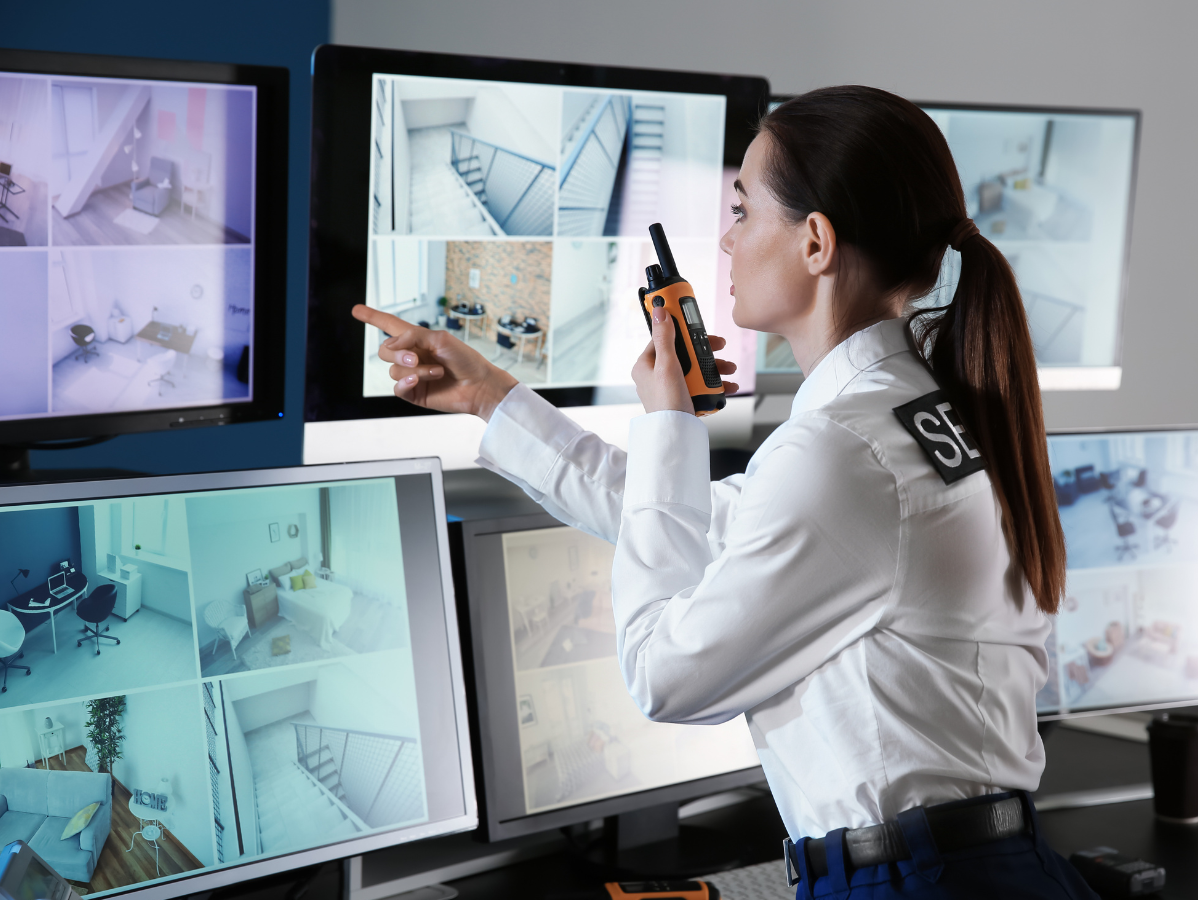 Security guard pointing at monitors displaying surveillance footage, holding a walkie-talkie.