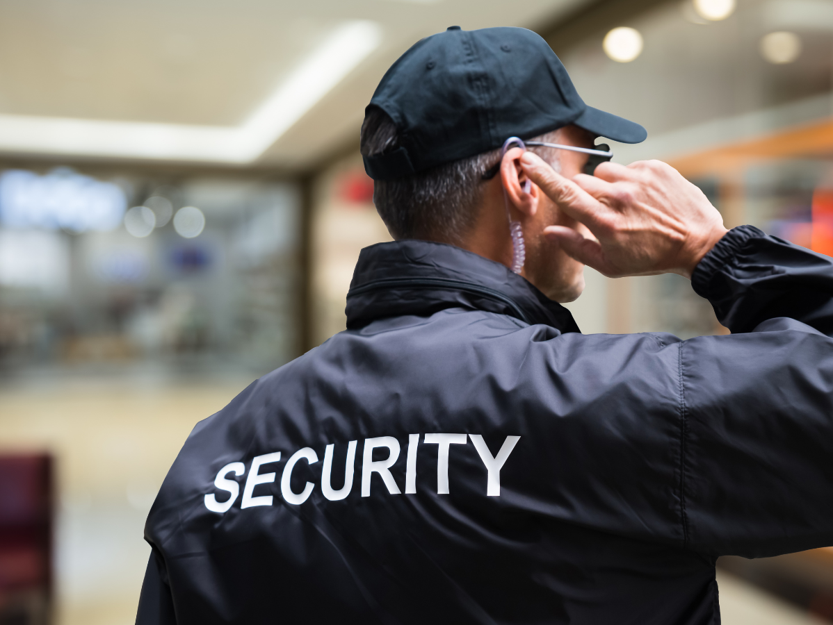 Security guard with earpiece, wearing a black jacket and cap, in a mall.