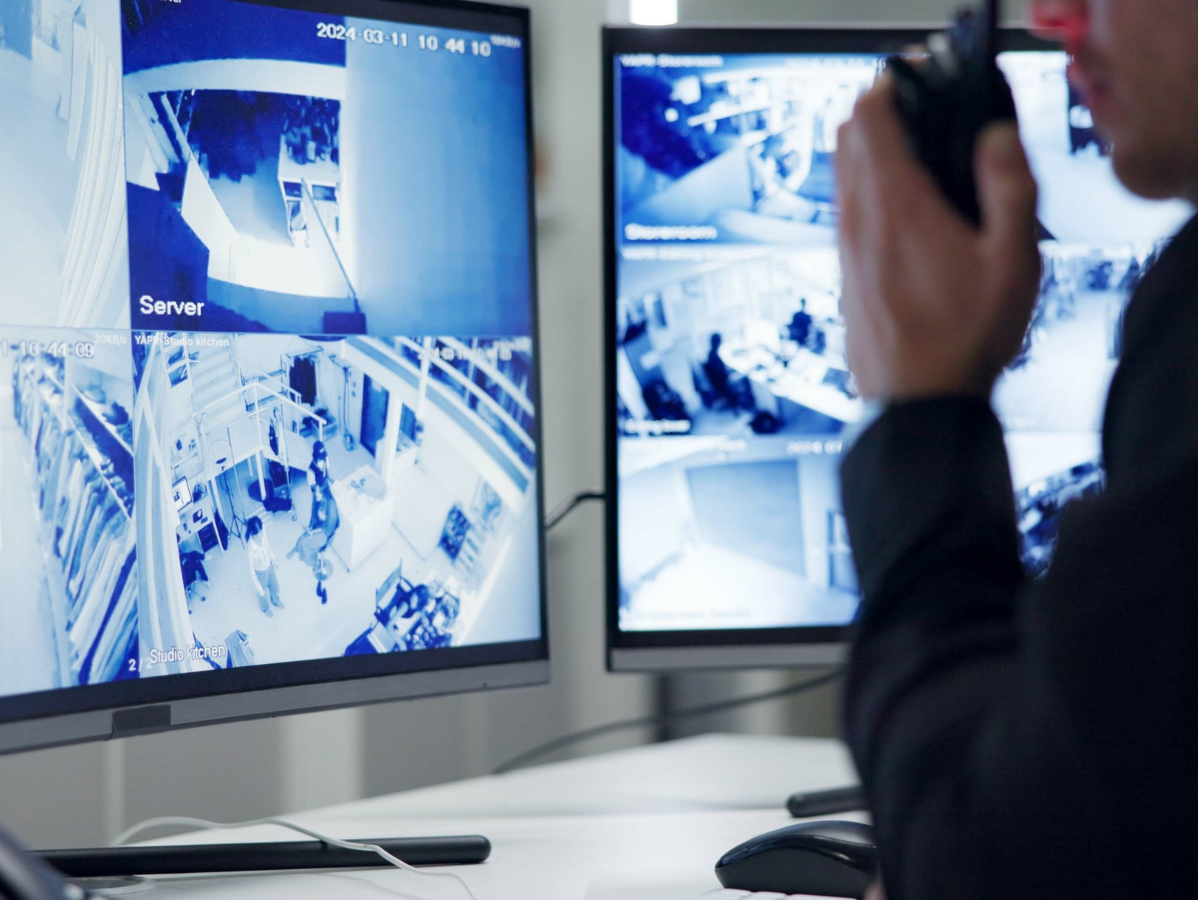 Security guard monitoring multiple surveillance camera feeds on a computer monitor; holding a walkie-talkie.
