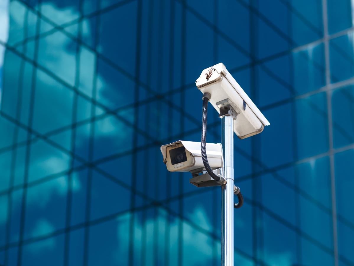 Two white security cameras on a pole, in front of a blue glass building.