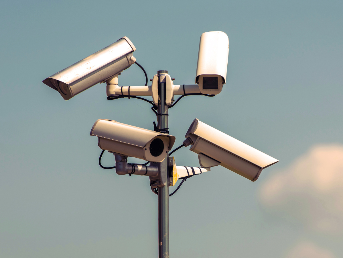 Four security cameras on a pole, aimed in different directions, against a light blue sky.