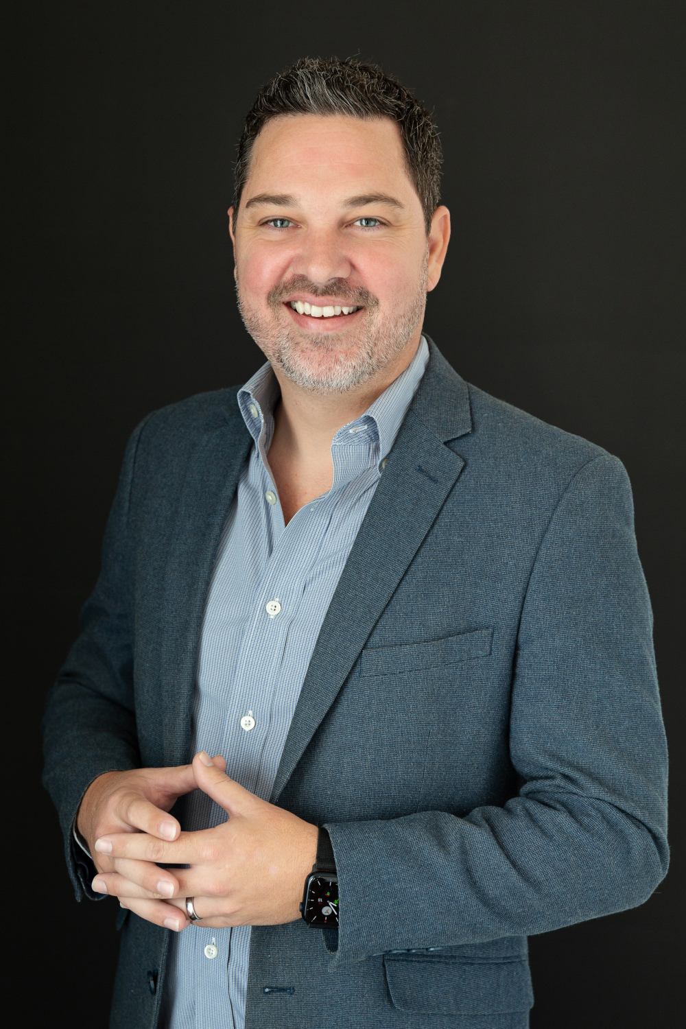 Man in a blue suit, white shirt, hands in pockets, standing in front of a modern home, green lawn.