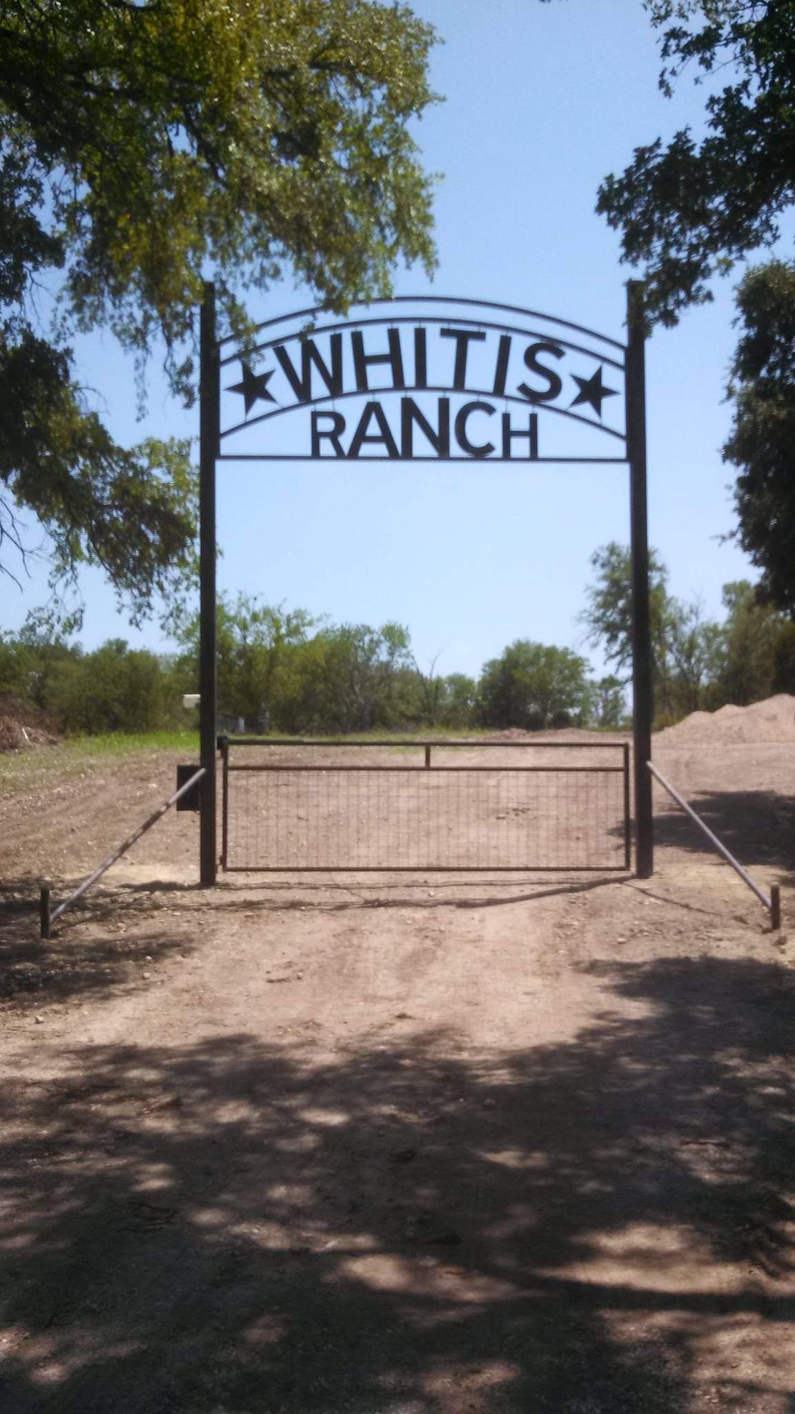 Black metal gate reading “WHAIT’S RANCH” at a sunny dirt entrance, with trees and a gravel road behind it.