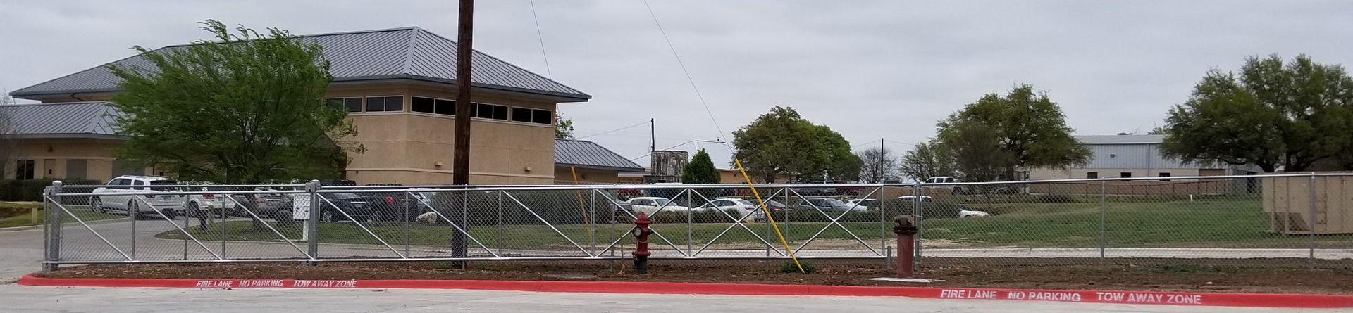 A low building behind a fenced roadside with parked cars, trees, and a cloudy sky.