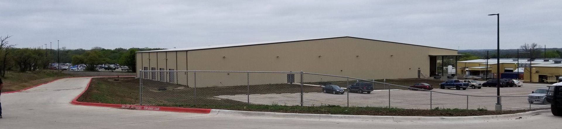Wide beige industrial warehouse beside a chain-link fence and parking lot under a cloudy sky