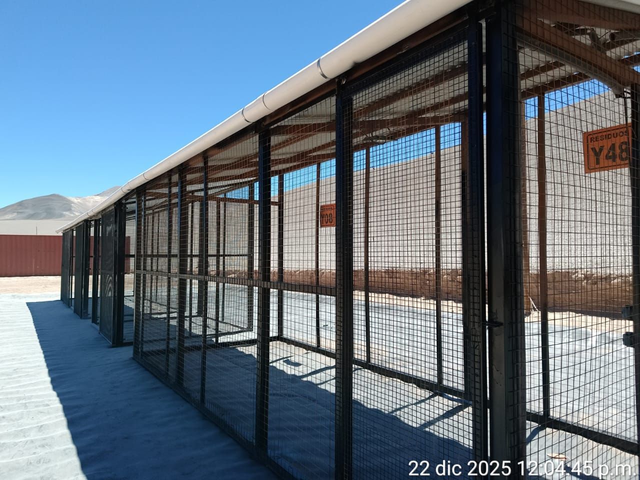 Larga hilera de jaulas metálicas negras con laterales de malla metálica, bajo un techo blanco. Las jaulas se encuentran en un entorno exterior soleado.