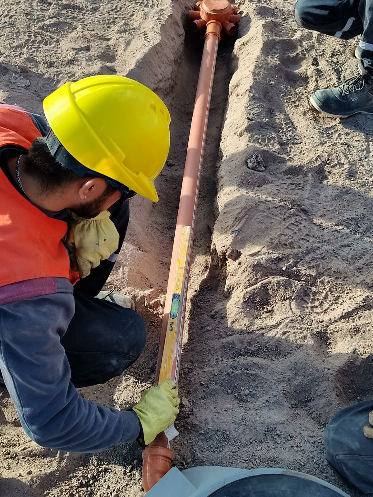Un trabajador de la construcción con un casco amarillo comprueba el nivel de una tubería en una zanja de arena.