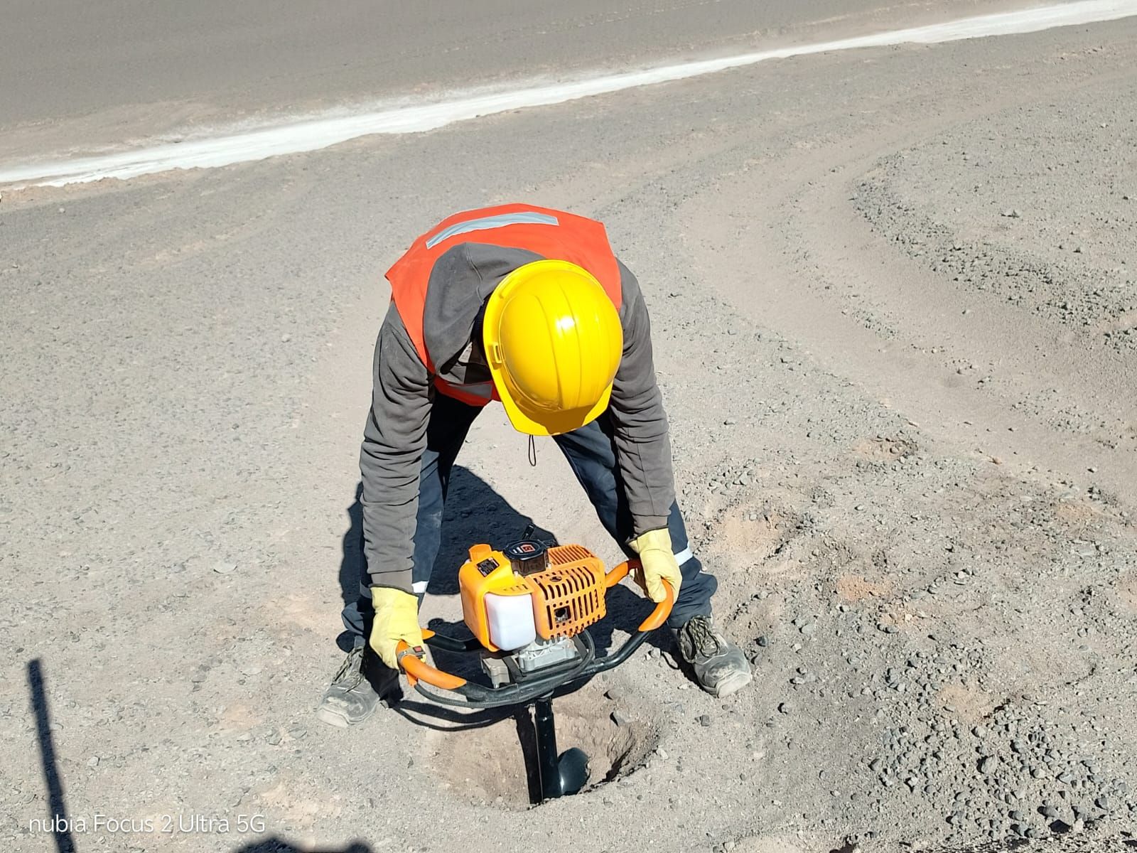 Trabajador de la construcción con casco amarillo utilizando un taladro en una carretera, vistiendo chaleco de seguridad y guantes.