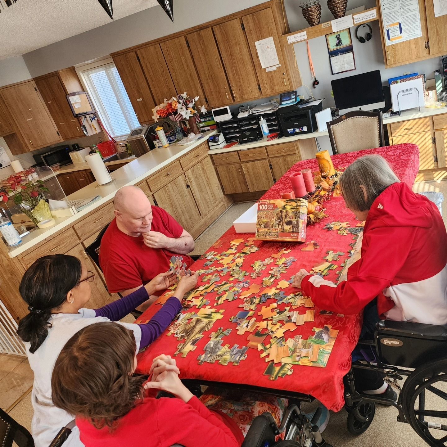 Four people seated around a table, working on a puzzle in a kitchen. One in a wheelchair, red table cover.