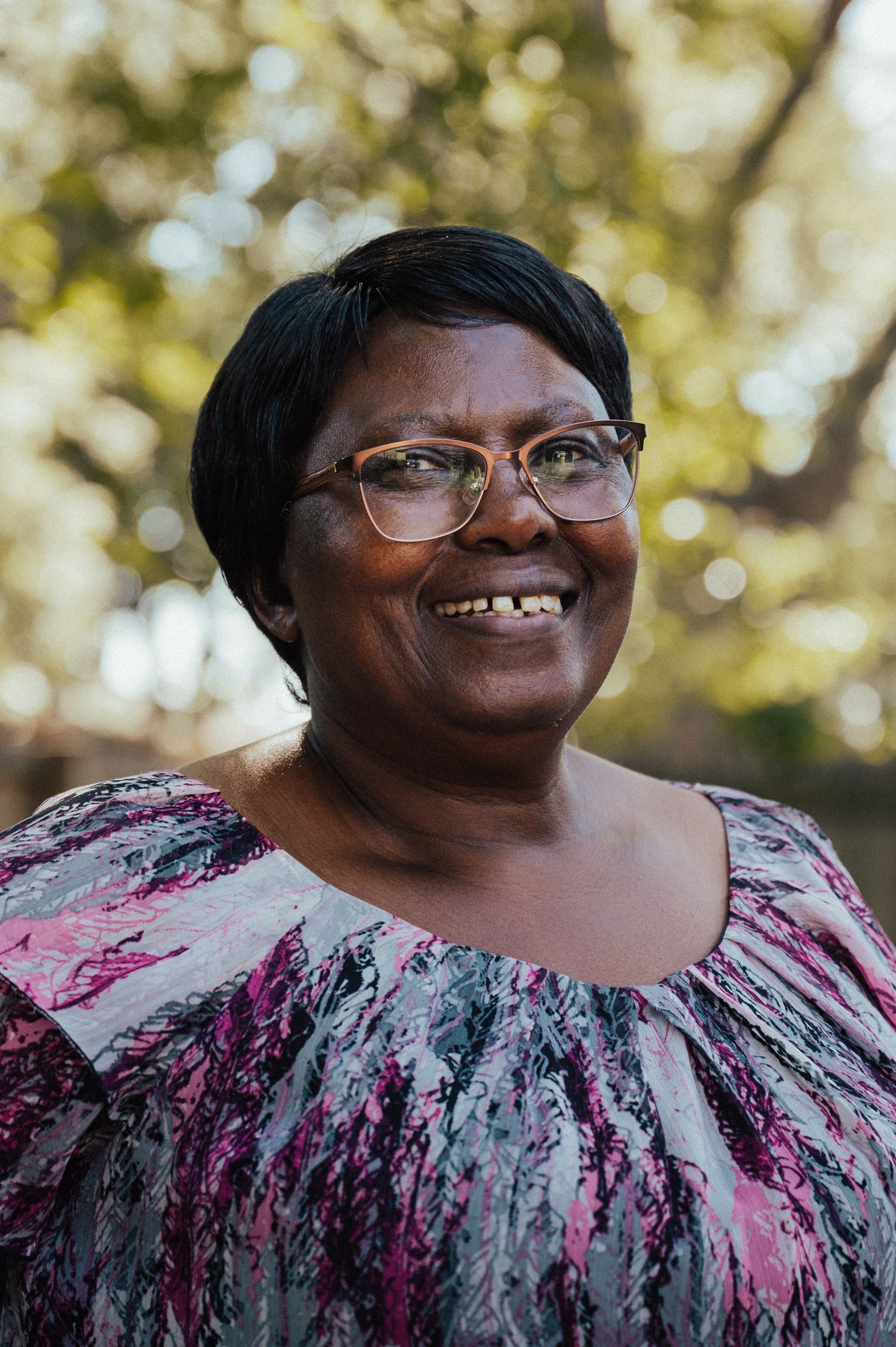 Smiling Black woman with glasses in a pink and purple top, outdoors in a sunny setting.
