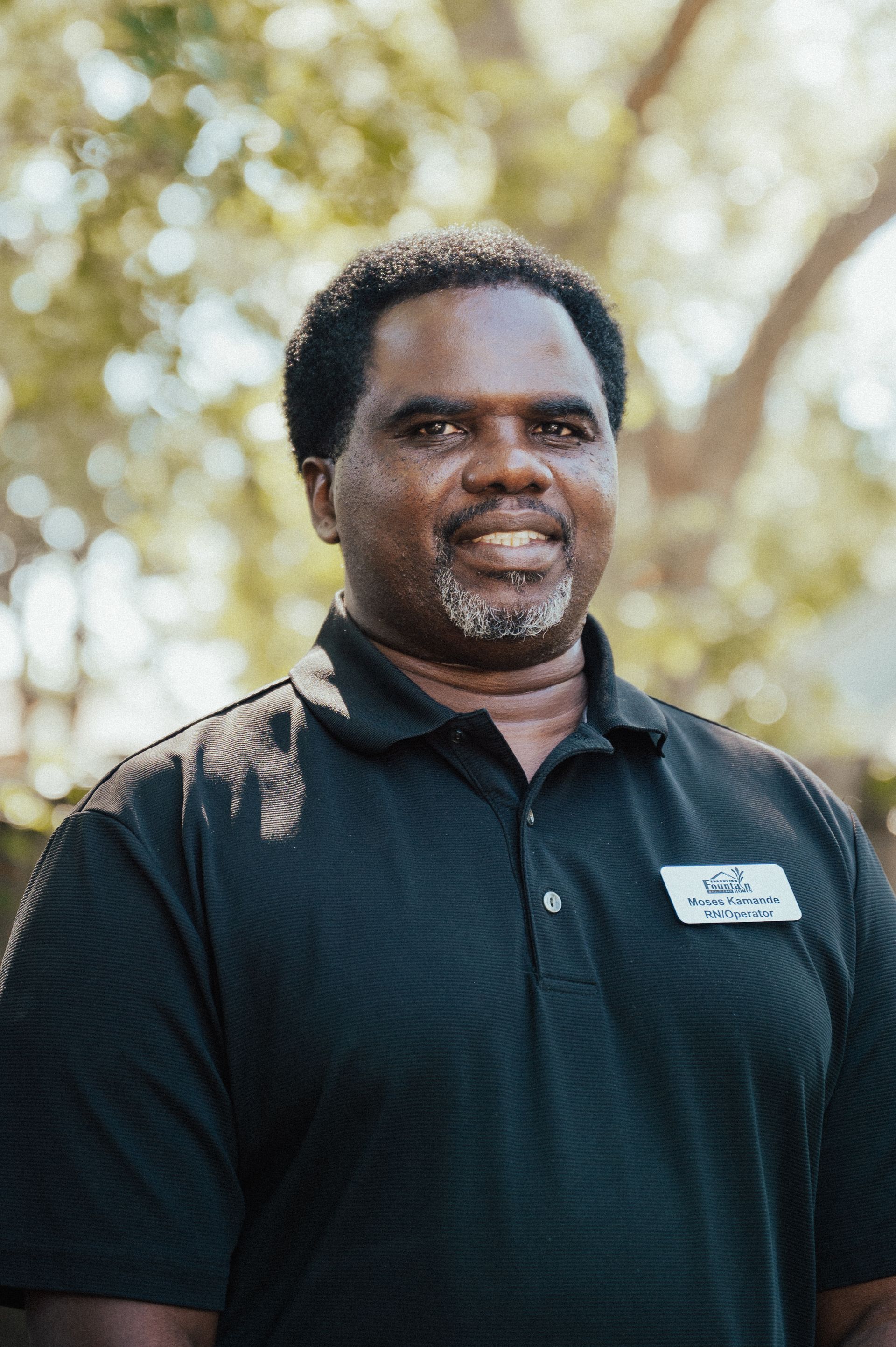 Smiling Black man in black polo shirt, outdoors, wearing a name tag.