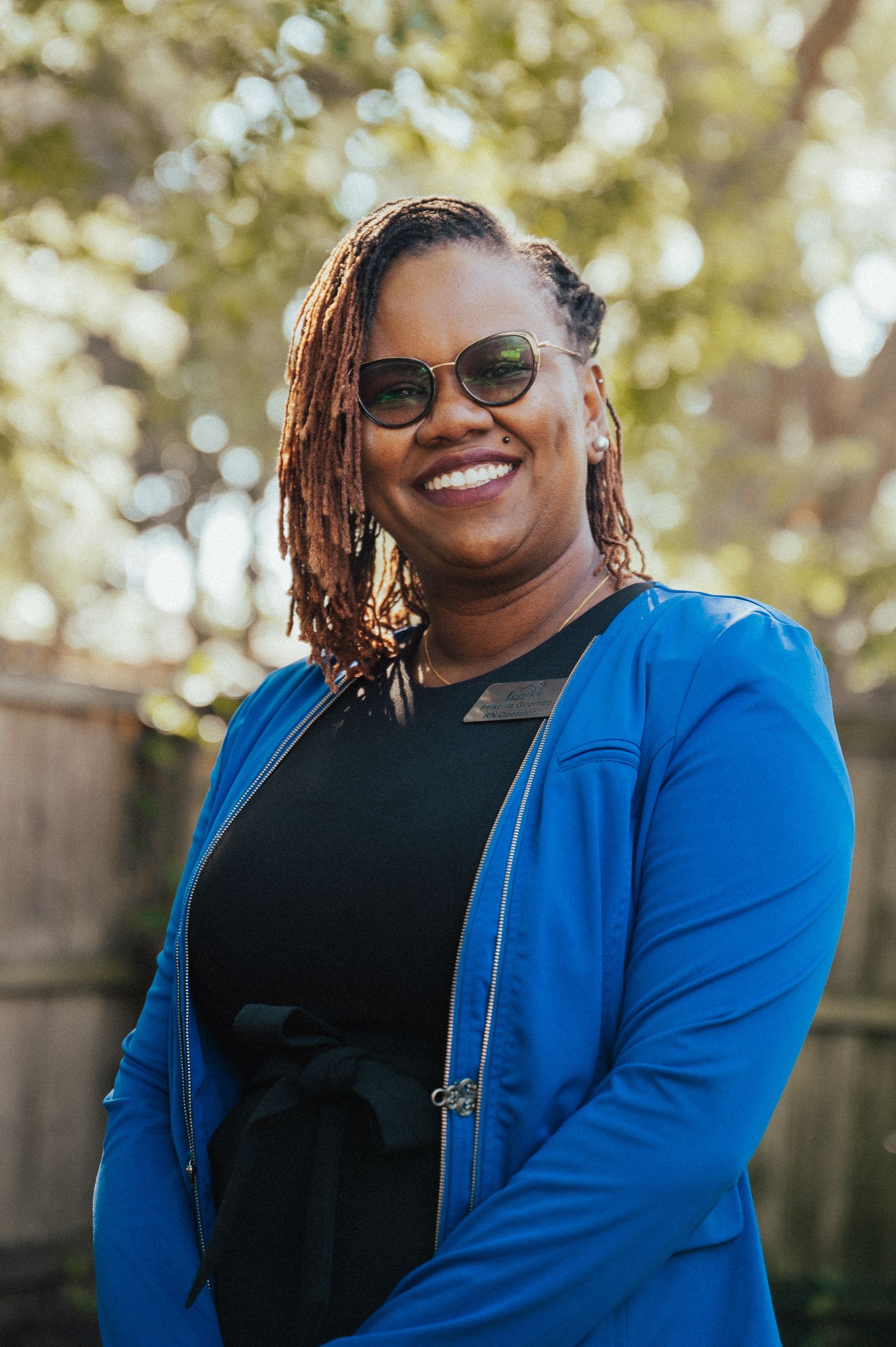 Woman with locs smiles wearing sunglasses and a blue blazer. Outdoors, near a fence.