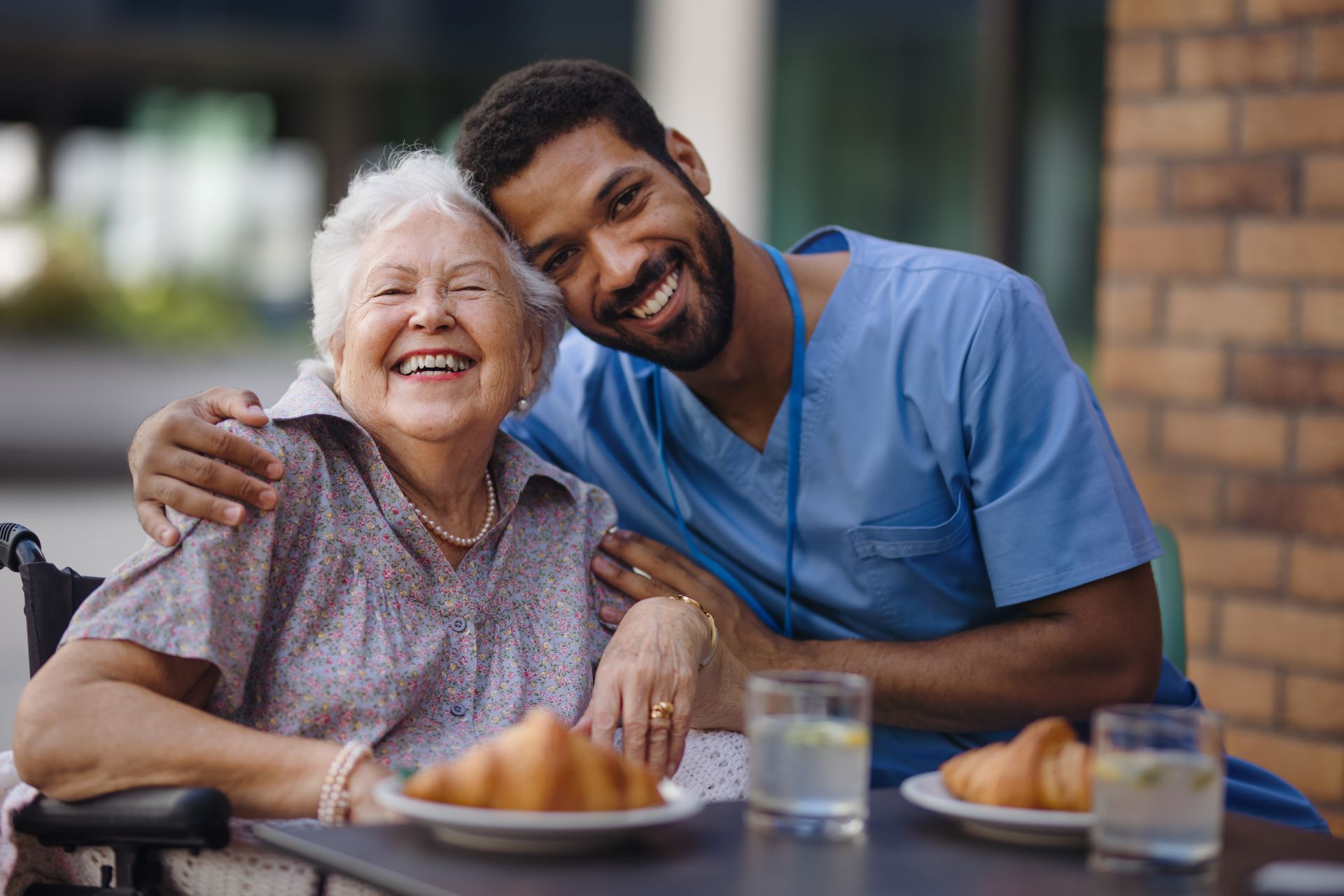 Smiling senior woman in wheelchair with arm around her by smiling male caregiver; outdoor cafe setting.