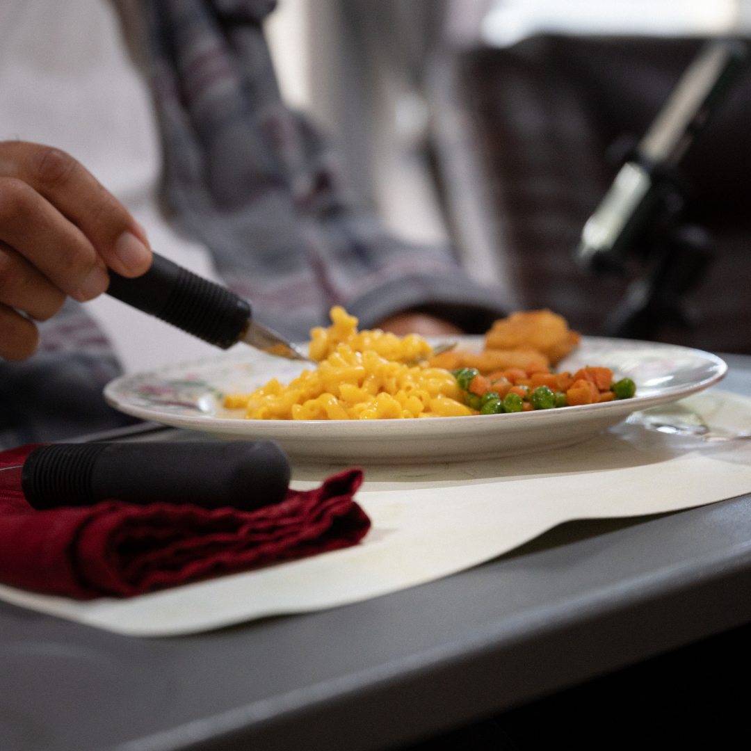Person using adaptive utensil to eat mac and cheese. Plate on a tray, with napkin.