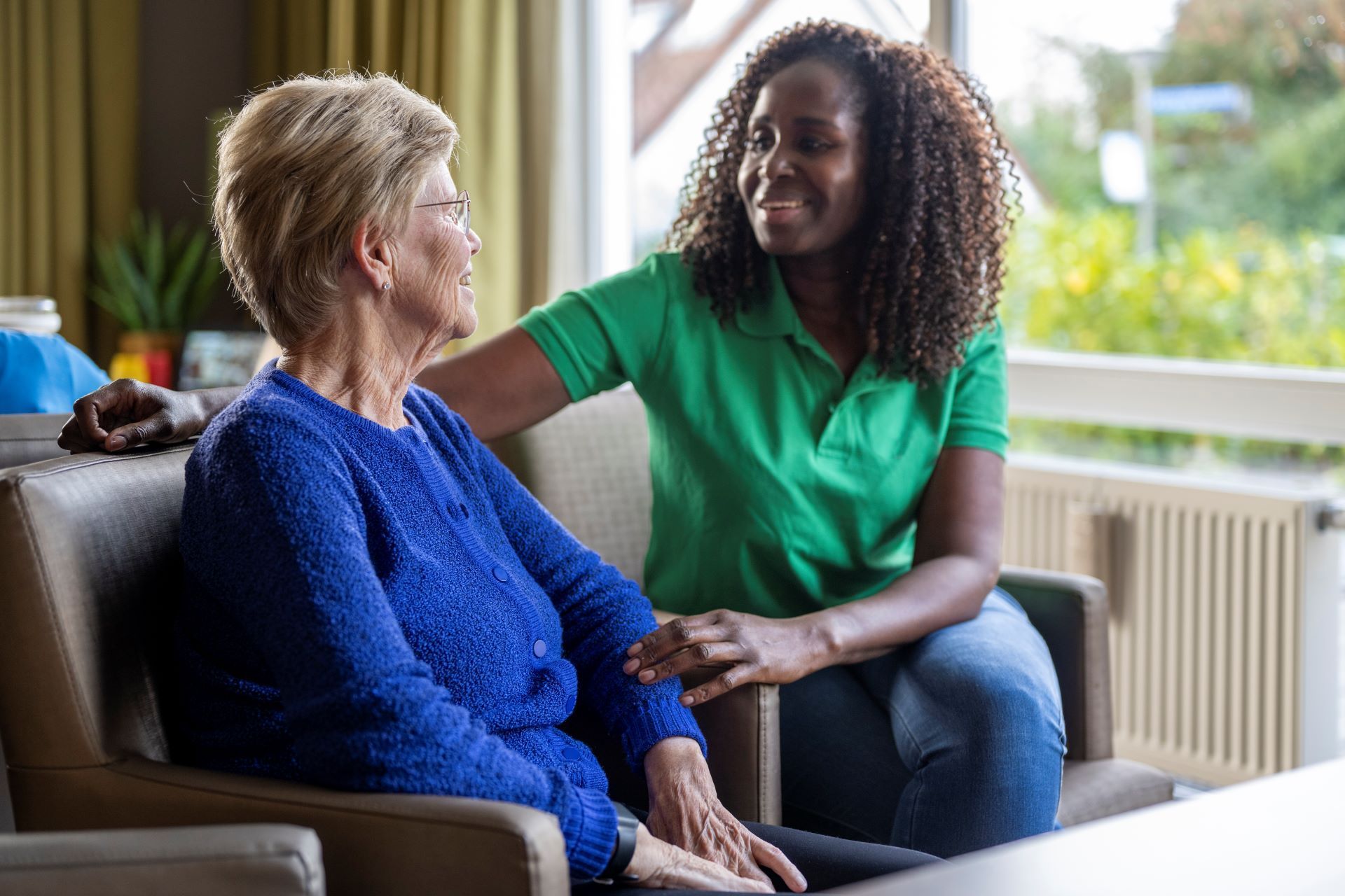 Caregiver comforts elderly person sitting indoors, green shirt, blue sweater.