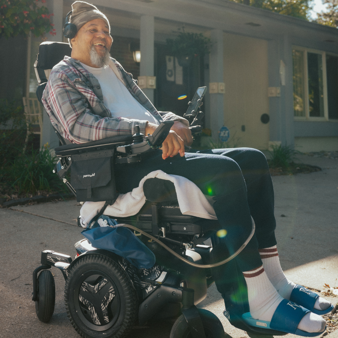 Man in wheelchair smiles, outdoors in front of a house, wearing a plaid shirt, beanie, and sandals.