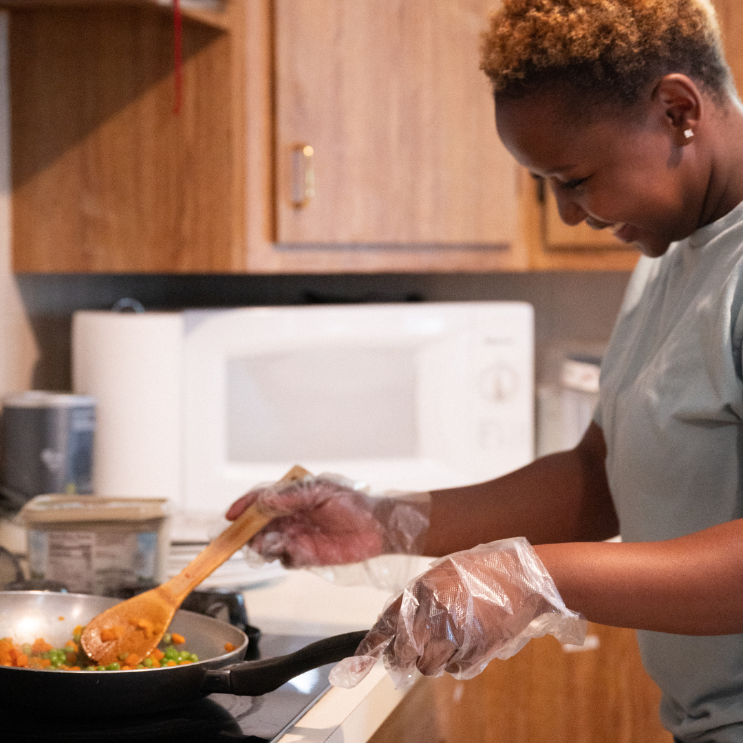 Woman in kitchen cooking, wearing gloves, stirring food in a pan. Microwave and cabinets visible.