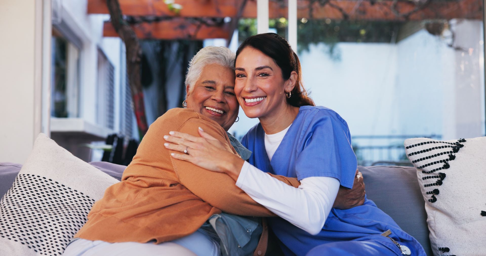 A caregiver in blue scrubs and a person in a tan cardigan hug warmly while sitting on a couch.
