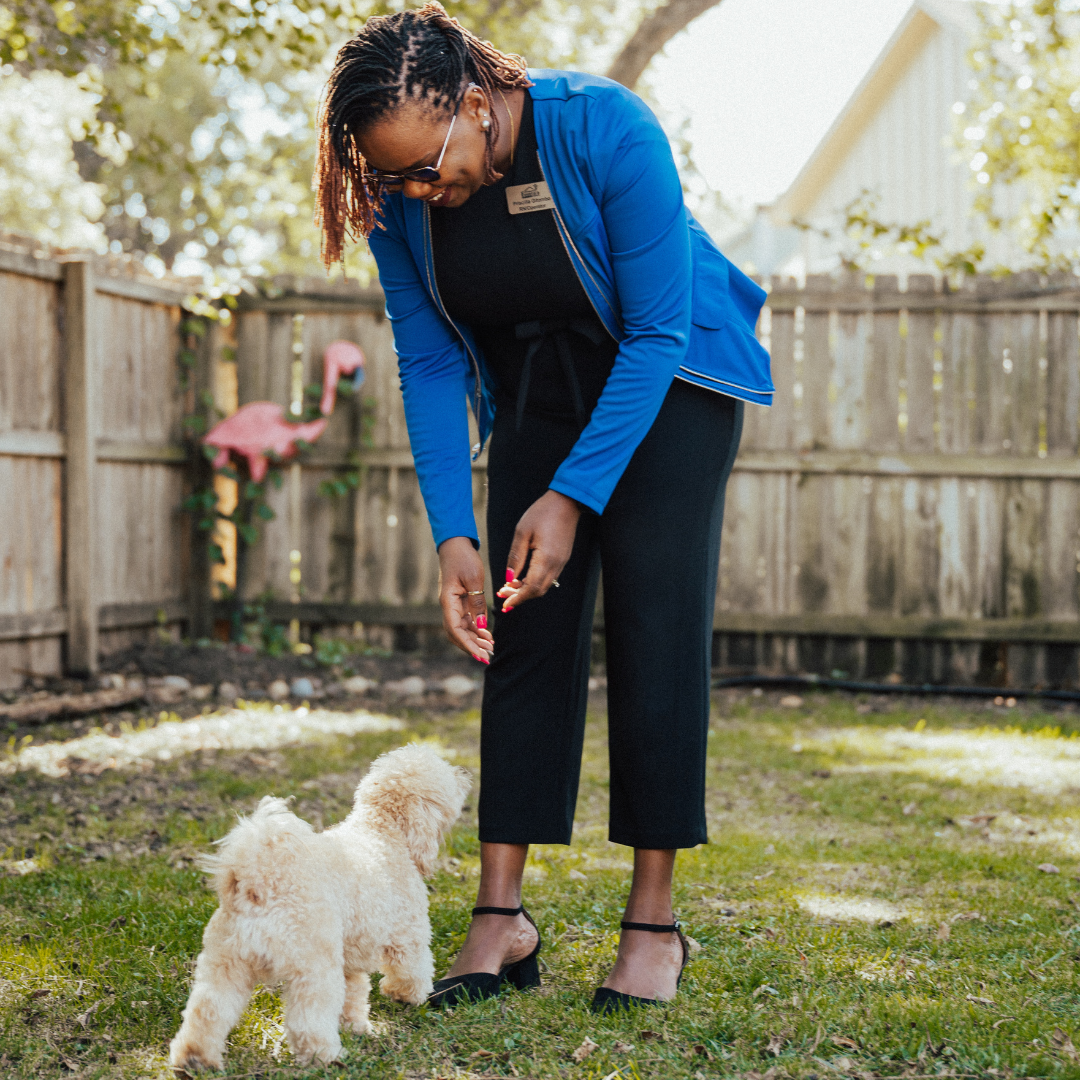 Woman in blue cardigan and black jumpsuit crouches to give a treat to a fluffy beige dog in a backyard.