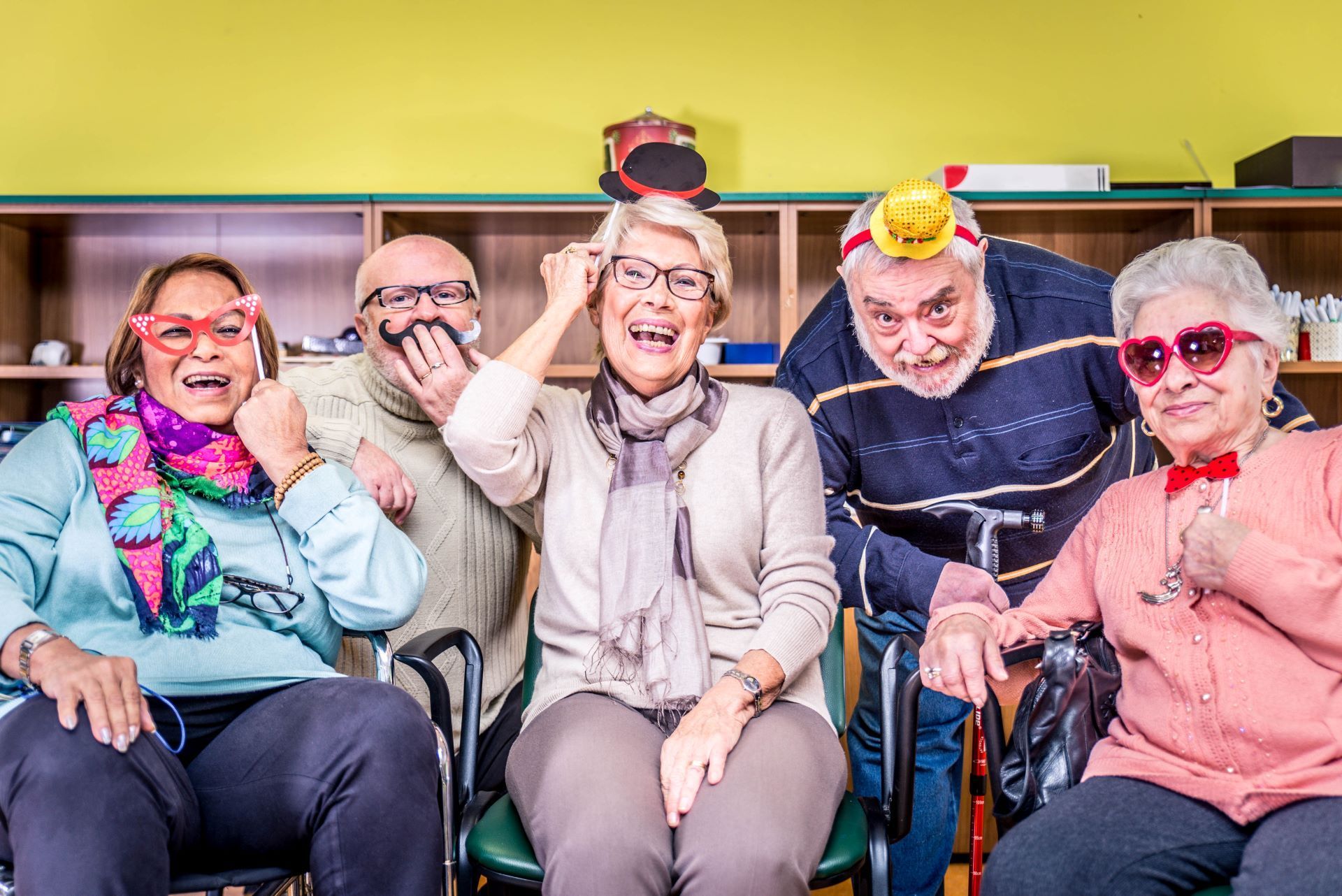 Five people in silly glasses and accessories smile at the camera in a room.