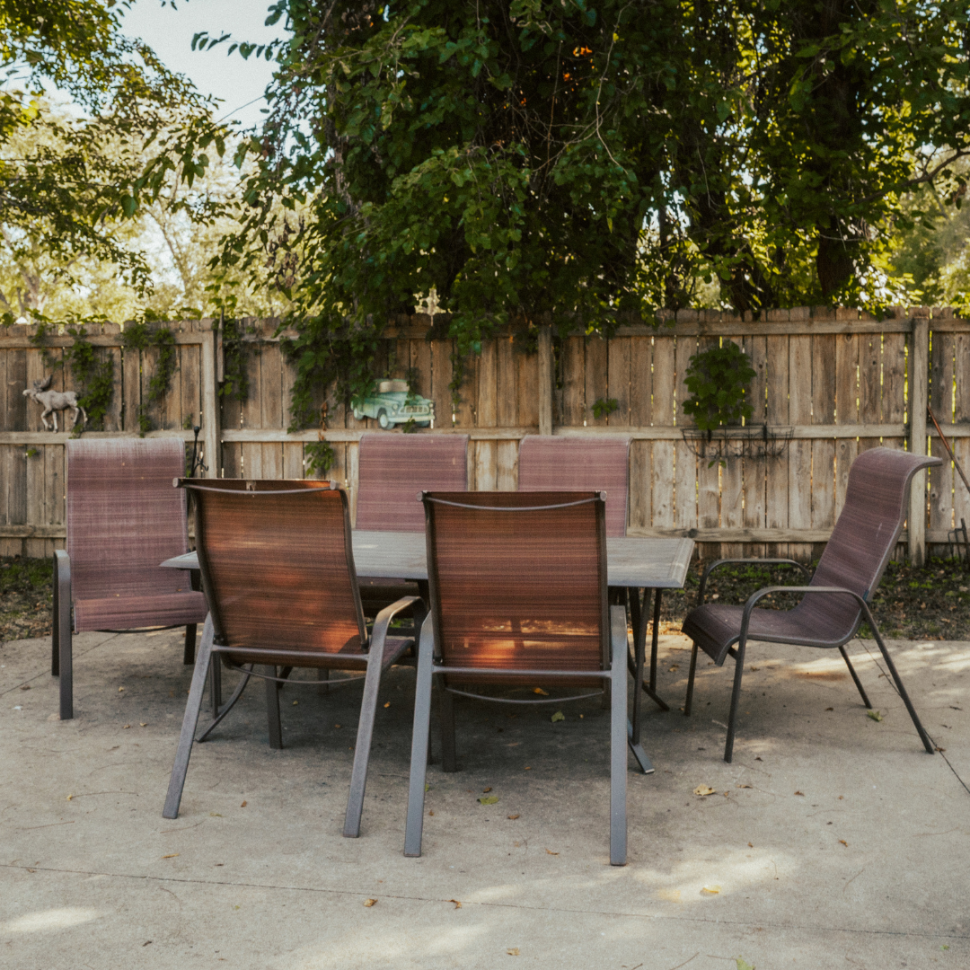 Patio table and chairs, set on concrete, against a wooden fence and trees.