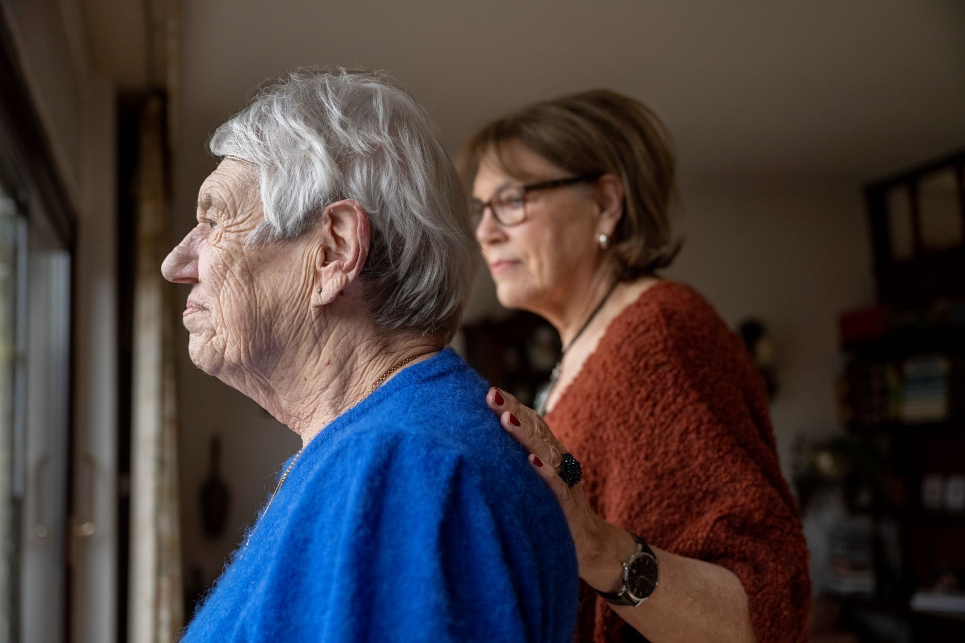 A woman is standing next to an elderly woman looking out of a window.