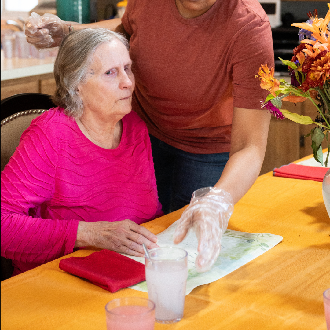 A caregiver assists an elderly woman at a table. The woman in pink looks toward her with a concerned expression.