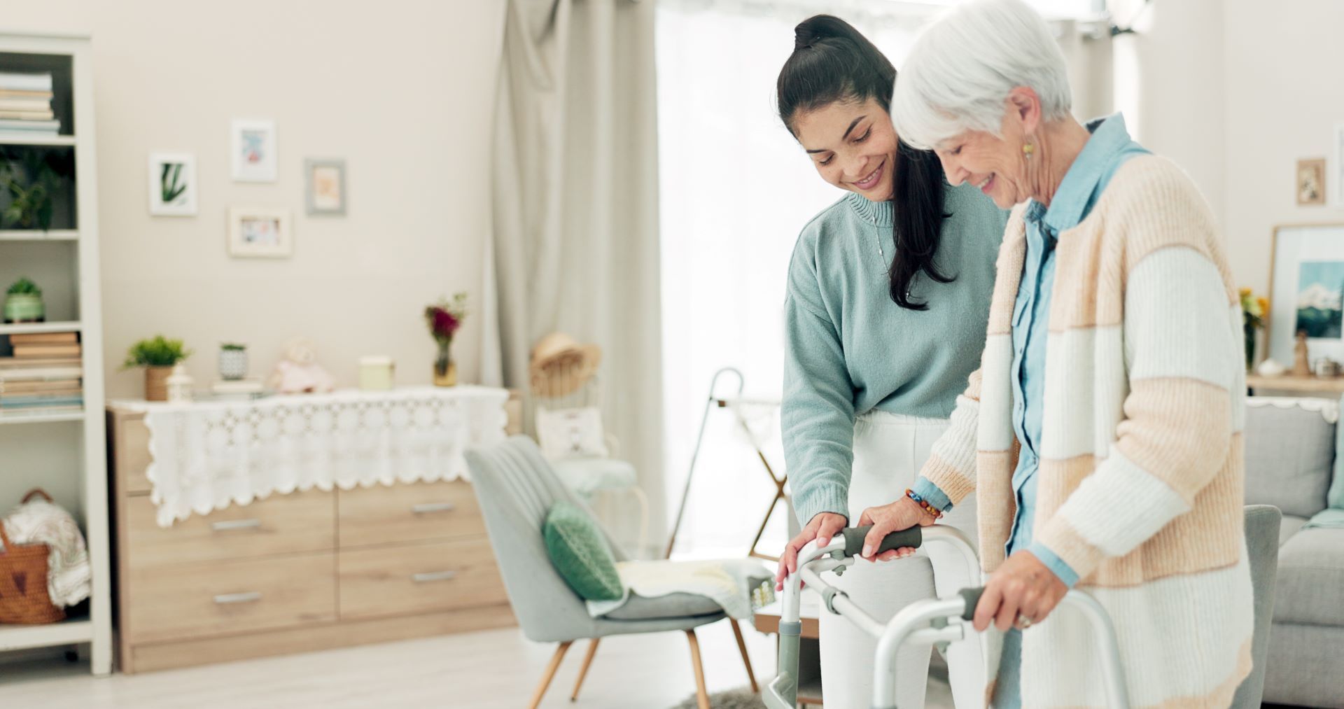 Young woman assisting an elderly woman with a walker in a living room; both smile.