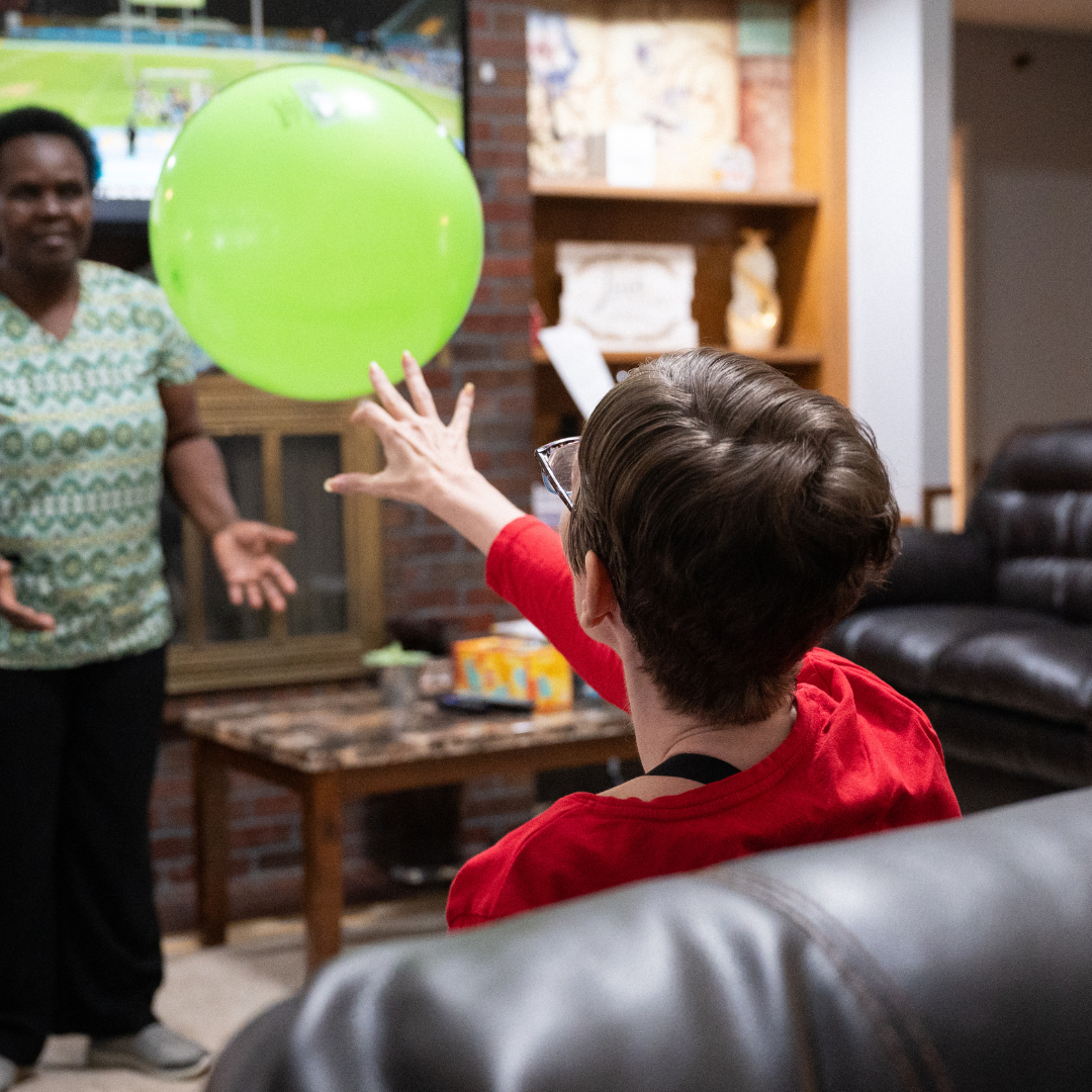 Two women playing with a green ball in a living room. One throws, the other prepares to catch.